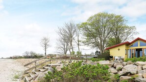 Une propriété située en bord de mer, avec un escalier en bois menant à un bâtiment jaune, entouré de gros rochers et d’une végétation clairsemée.