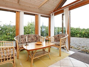Une véranda avec une table en bois et deux chaises en osier, de grandes fenêtres et une vue sur la verdure.