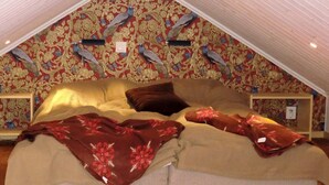 Une chambre avec un plafond en pente, un papier peint à motifs floraux, un lit recouvert d’une couverture à fleurs et une table de chevet avec une lampe.