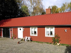 En röd byggnad med en grusuppfart, en vit dörr och två fönster med gardiner.