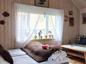 Un dormitorio con cama, una ventana con cortinas, una mesita de noche con lámpara y un pequeño estante con objetos decorativos.