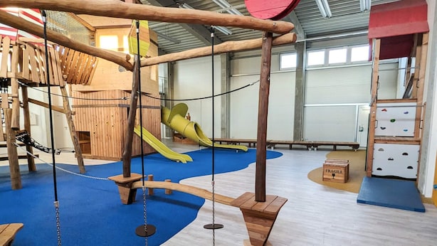 An indoor play area with wooden climbing structures, a slide, and a padded floor.