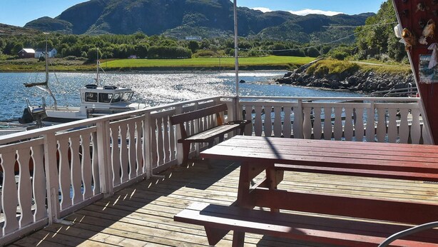 A wooden deck with a bench and table overlooking a body of water with boats and mountains in the background. - 6 Person Holiday Home in Oksvoll (Orland)
