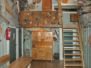 Een rustieke kamer met een houten bank, een ladder, een houten kast en diverse gereedschappen en objecten aan de muren.