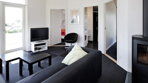 A modern living room with a black sofa, a television on a white stand, a fireplace, and a view through a large window. - 5 Star Holiday Home in Haderslev (Haderslev)
