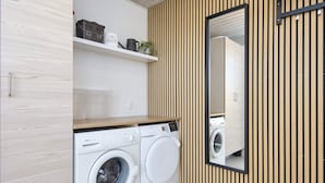 A compact laundry room with two white front-loading washing machines, a wooden slatted wall, a mirror, and a shelf with laundry items. - Spacious Holiday Home in Hemmet Jutland near Sea (Hemmet)