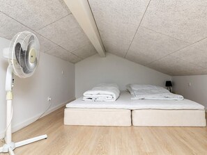 Une pièce avec un plafond en pente, un lit avec deux oreillers, un ventilateur sur pied et une table de chevet avec une lampe.