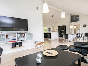 A modern living room with a flat-screen TV, bookshelves, a dining area with a table and chairs, and a kitchen in the background.
