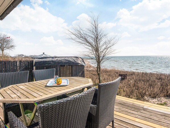 A wooden deck with wicker chairs and a table, overlooking a beach and the sea.