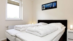 A double bed with white bedding, a nightstand with a lamp, a window with blinds, and a framed picture of lighthouses on the wall. - Holiday Home in GroÃŸenbrode (Grossenbrode)