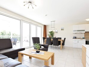 Un salon moderne comprenant un canapé, une table à manger avec des chaises, un coin cuisine avec des placards et une cuisinière, ainsi que de grandes fenêtres donnant sur l’extérieur.