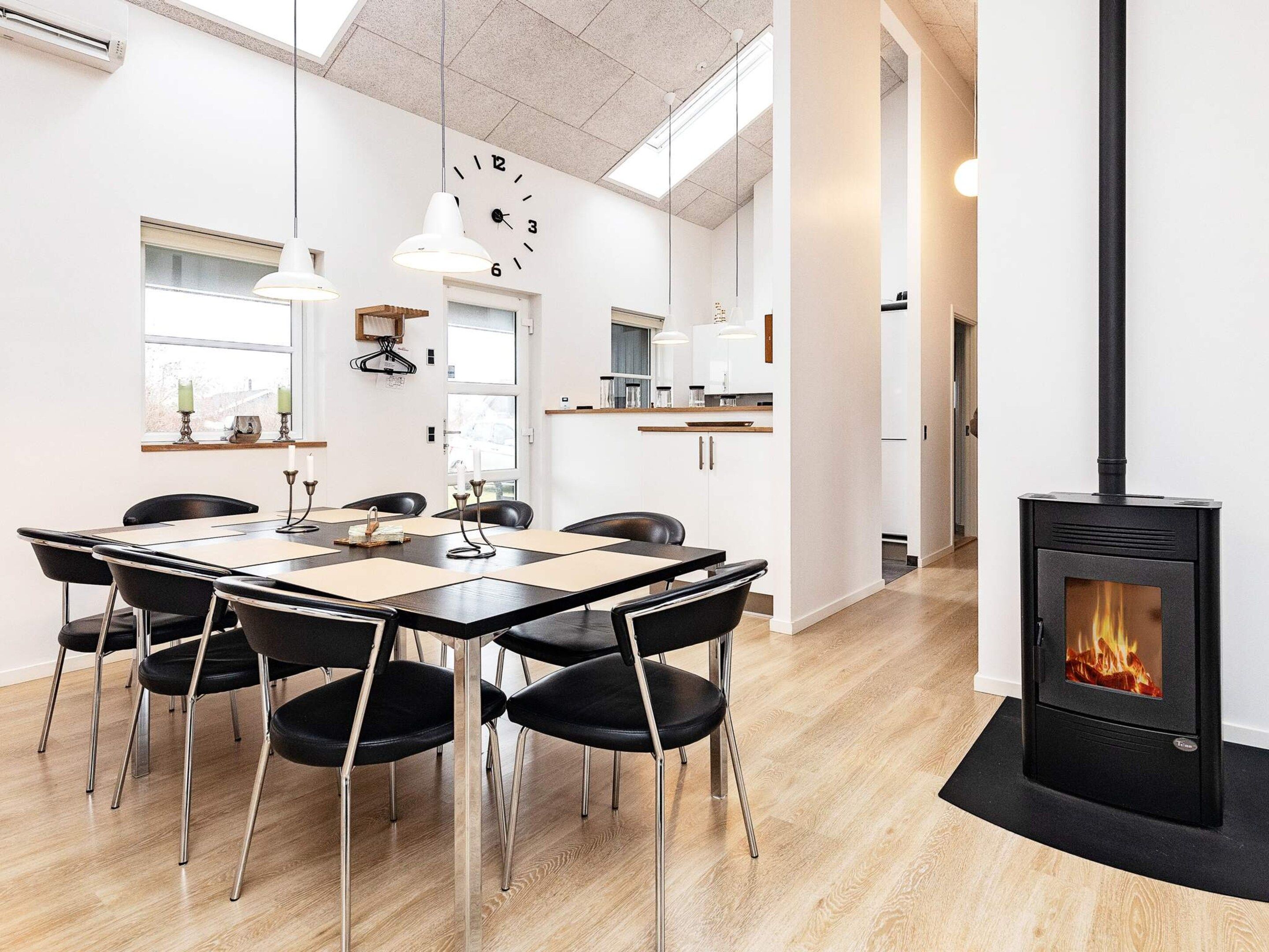 A modern dining area with a wooden table and chairs, a black stove with a visible fire, and a kitchen in the background.