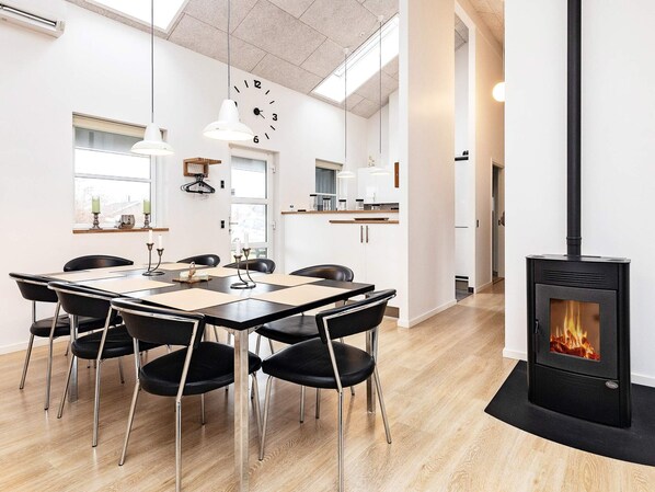 A modern dining area with a wooden table and chairs, a black stove with a visible fire, and a kitchen in the background.