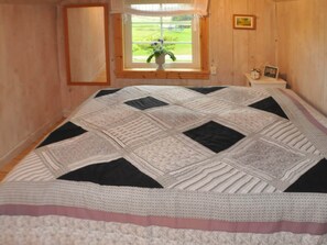 A bedroom with a large bed, a window with a view of greenery, a bedside table with a clock and picture, and wooden walls. - Holiday Home in Mariestad (Mariestad)