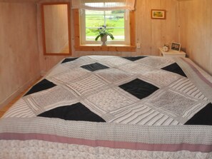 Een slaapkamer met een groot bed, een raam met uitzicht op groen, een nachtkastje met een klok en foto, en houten wanden.
