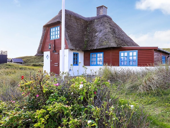 A traditional thatched-roof house with a white and red exterior, situated in a natural landscape with wildflowers.