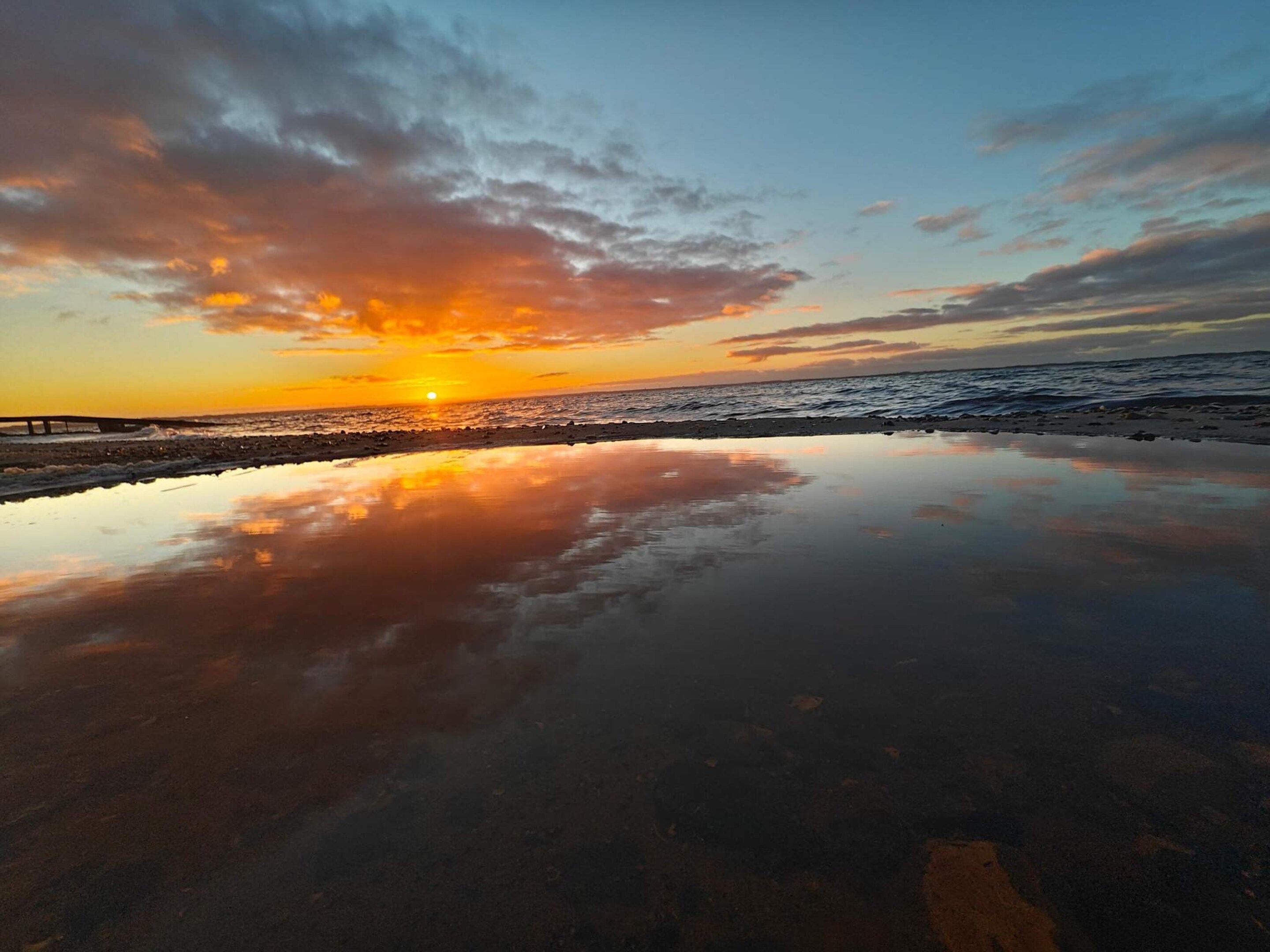 A beach with a sunset, calm water, and a pier in the background.