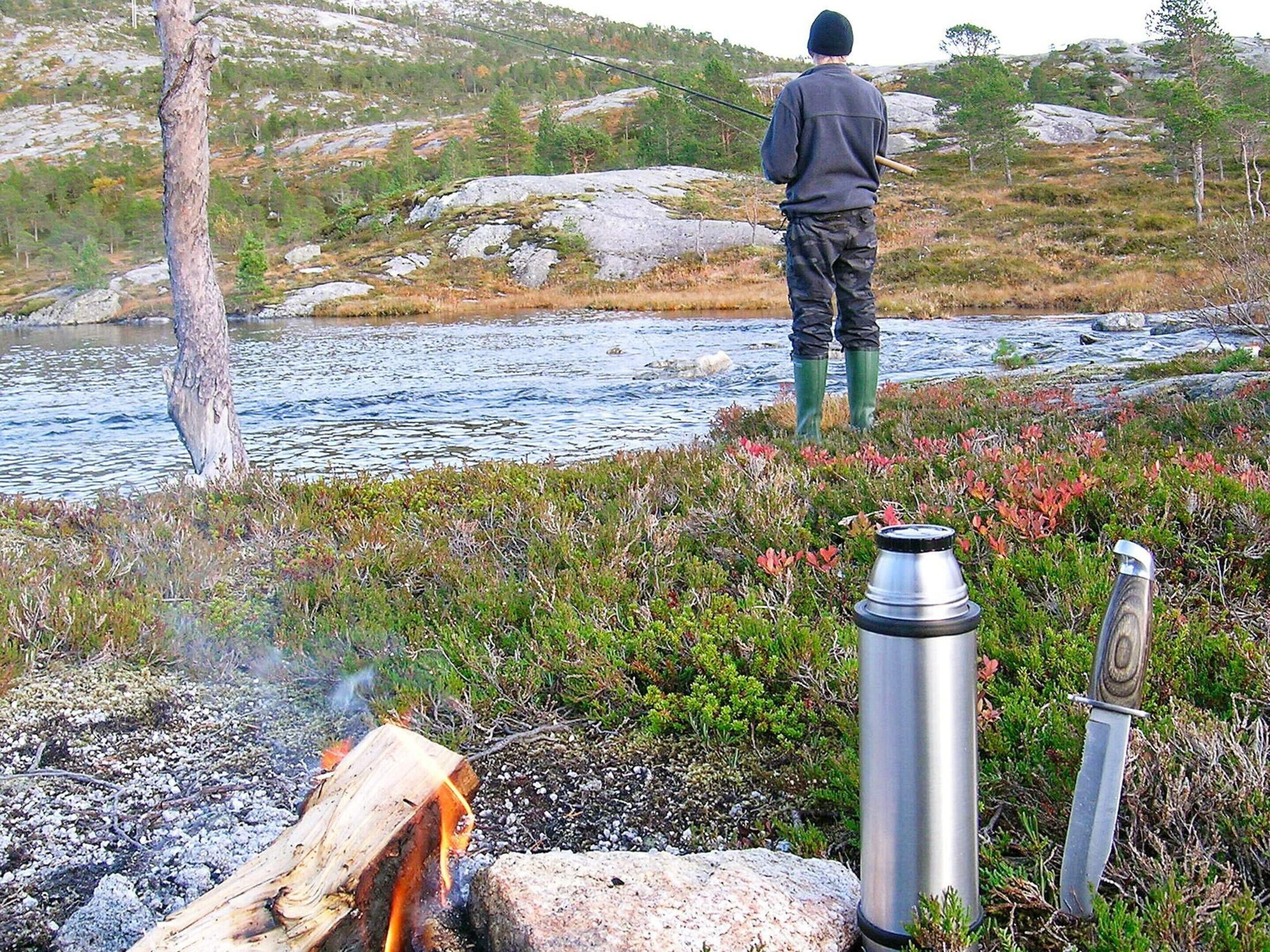 Una persona pesca in riva a un fiume, con un fuoco di campo e un thermos a terra.
