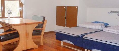 A room with a wooden dining table, a single chair, a bed with two mattresses, a wardrobe, and a window with curtains.