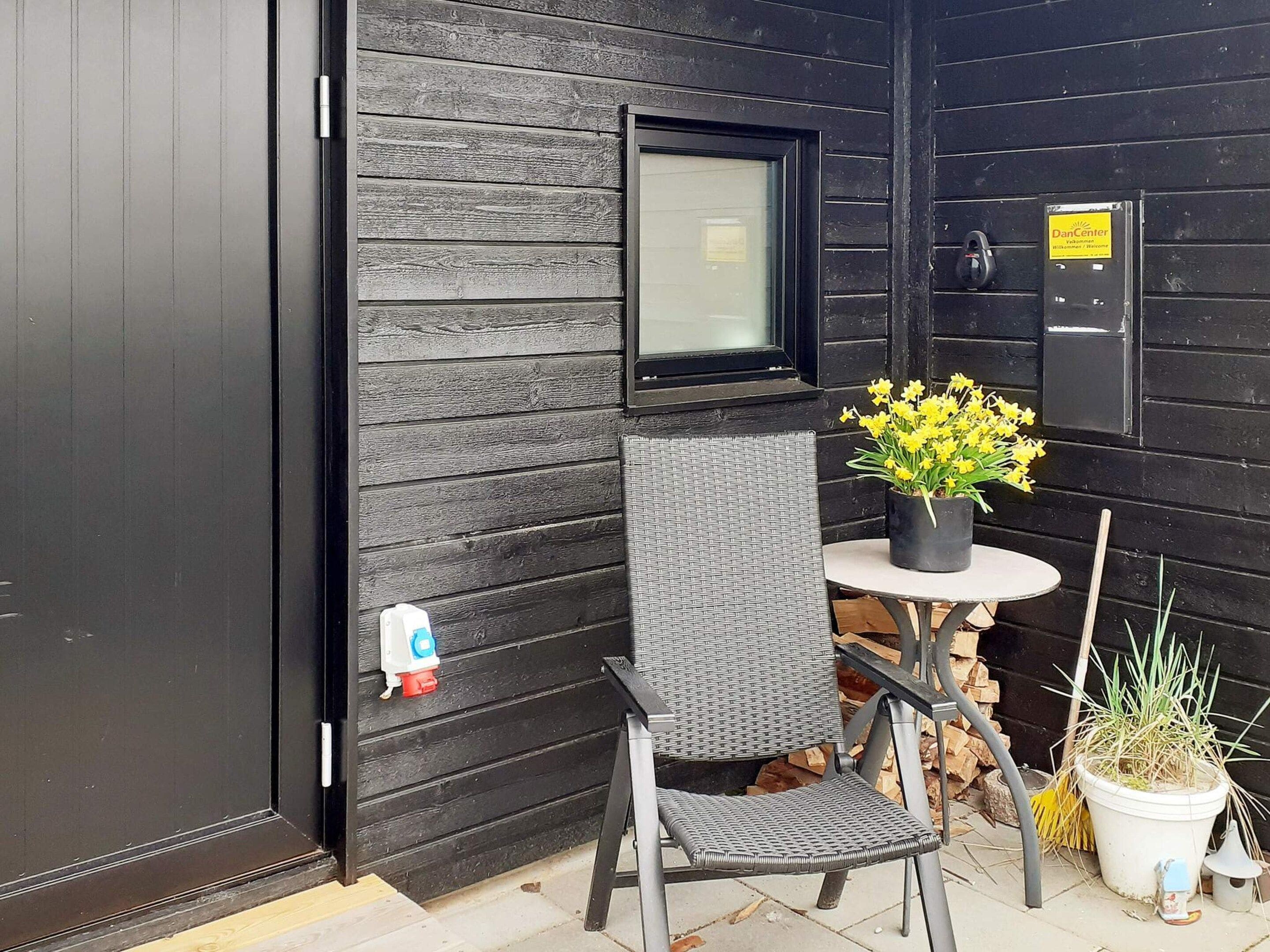 Eine schwarze Holzausfachung mit einer Tür, einem Fenster, einem Stuhl, einem kleinen runden Tisch und einem Blumentopf mit gelben Blüten.