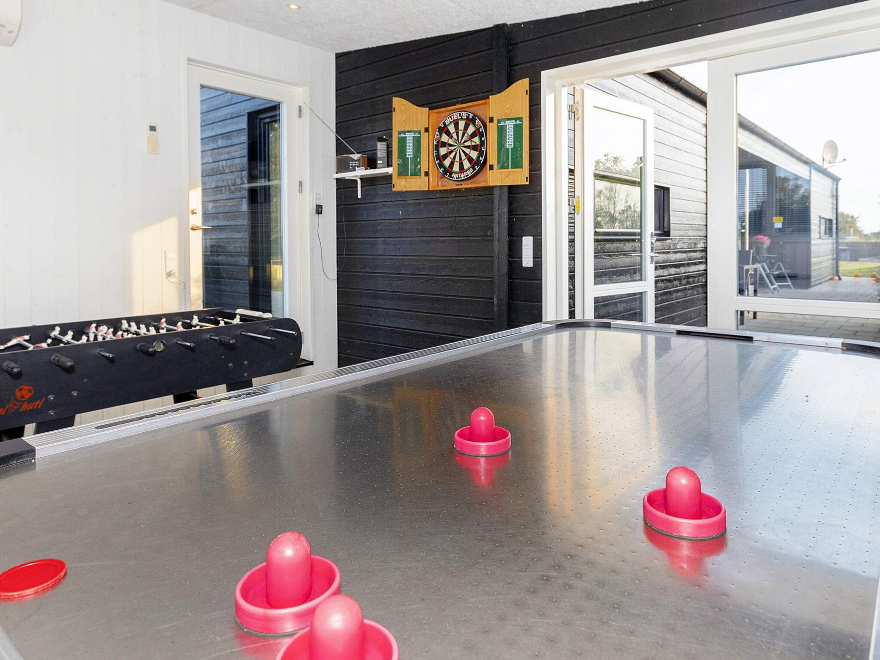 A foosball table with a dartboard and a pool table in a room with a glass door leading outside.