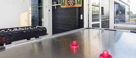 Une table de baby-foot, un jeu de fléchettes et une table de billard se trouvent dans une pièce dont la porte est en verre et donne sur l’extérieur.