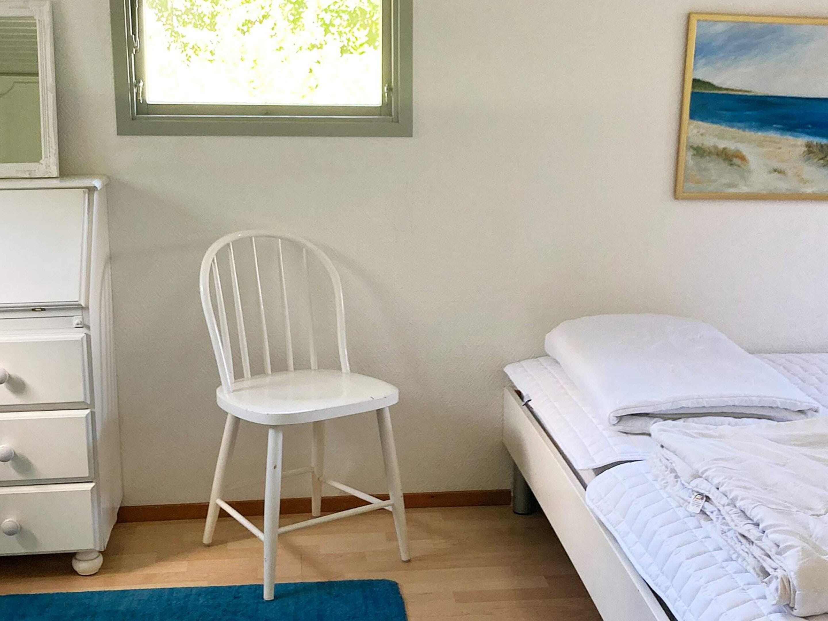 Une chambre à coucher avec un lit, une chaise blanche, une commode et une fenêtre donnant sur des arbres.