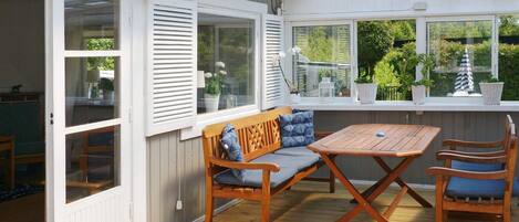 An outdoor patio with a wooden table and chairs, a bench with cushions, and a view of greenery through large windows.