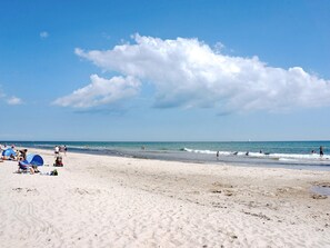 Una platja amb gent, alguns asseguts en cadiretes, i un veler a la distància.