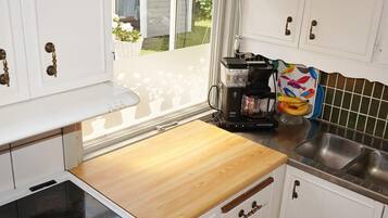 Cocina compacta con armarios blancos, encimera de madera, cafetera y ventana con vistas a una casa y vegetación.