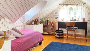 Une chambre mansardée chaleureuse, avec un plafond en pente, un lit recouvert d’une couverture rose, un bureau avec une chaise et une bibliothèque remplie de livres.