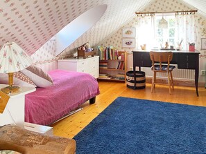 Un dormitorio acogedor en el ático, con techo inclinado, cama con manta rosa, escritorio con silla y estantería llena de libros.