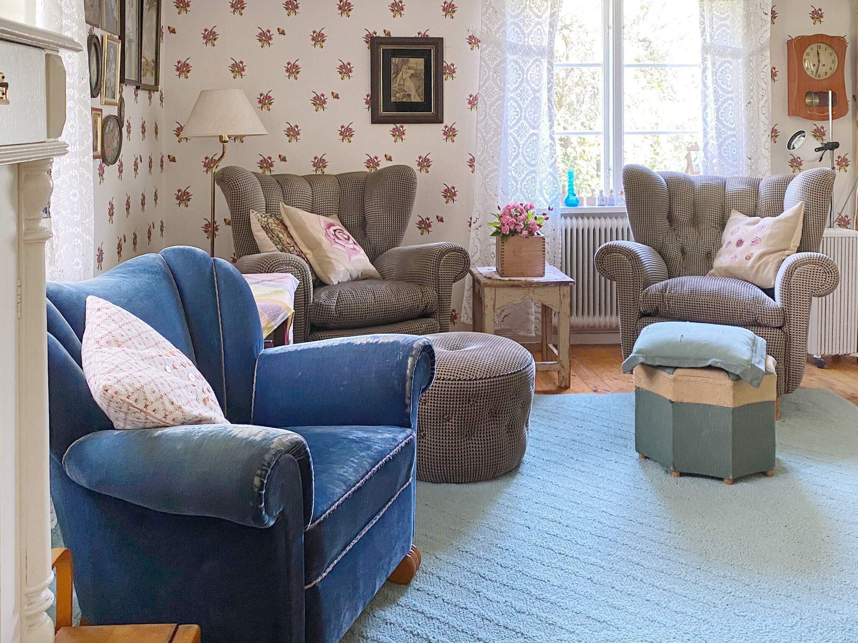 Ein gemütliches Wohnzimmer mit einem blauen Sessel, zwei gemusterten Sesseln, einem kleinen Tisch mit Blumenvase und einer Heizung.