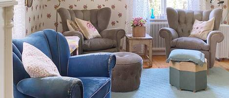 Ein gemütliches Wohnzimmer mit einem blauen Sessel, zwei gemusterten Sesseln, einem kleinen Tisch mit Blumenvase und einer Heizung.