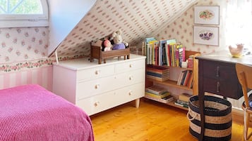 A cozy room with a sloped ceiling, a bed with a pink blanket, a desk with books, and a small shelf with decorative items.