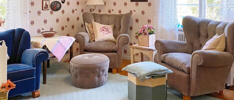 A cozy living room with patterned wallpaper, a blue armchair, a brown armchair, a matching ottoman, and a small side table with a lamp and flowers.