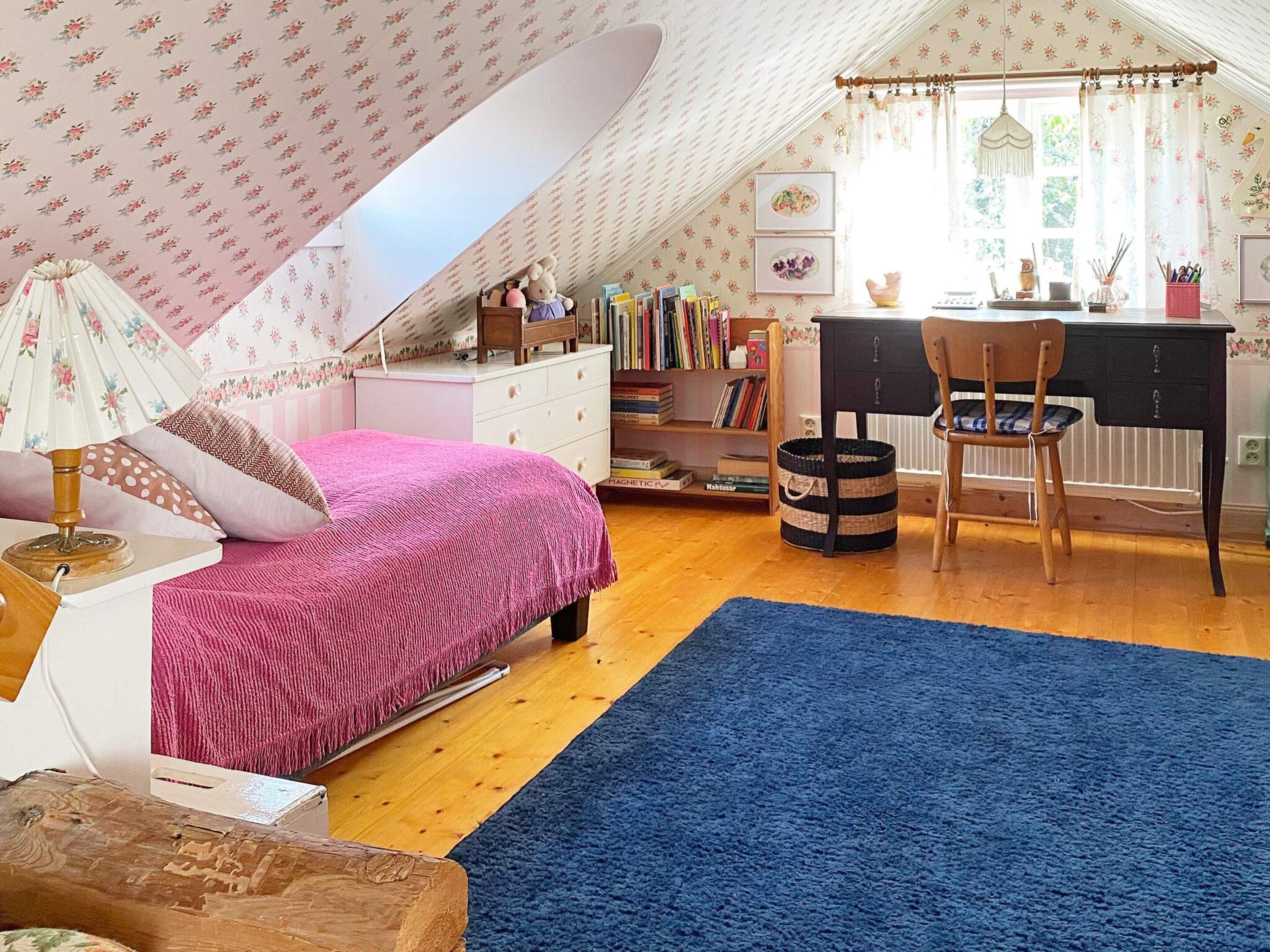 A cozy attic bedroom with a sloped ceiling, a bed with pink bedding, a desk with a chair, and a bookshelf filled with books.