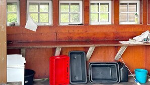 A room with wooden walls, a window with a white frame, and a shelf with various items including a red crate, black containers, and a blue bucket.