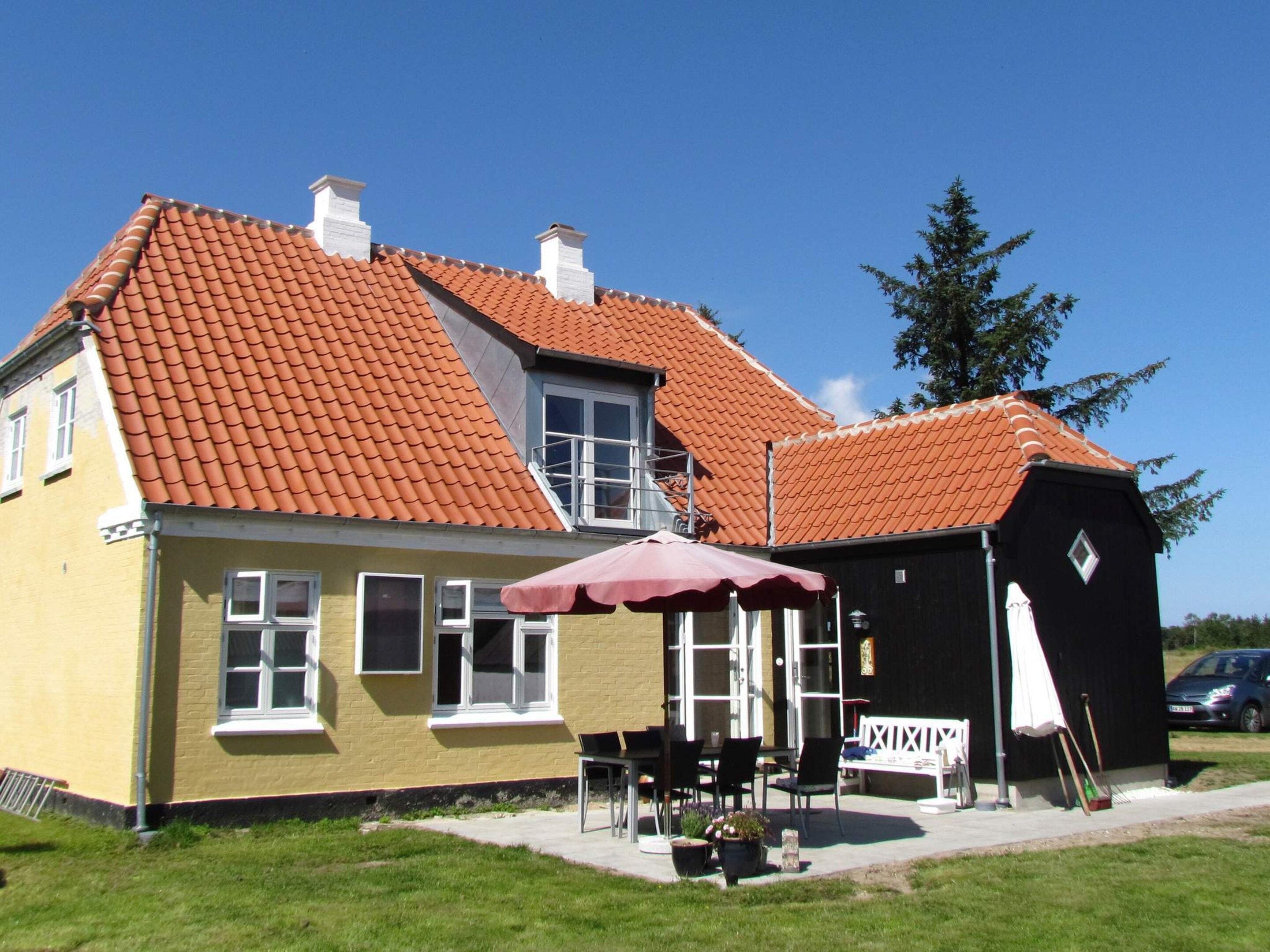 En gul bygning med rødt tegltag, et overdækket udendørs siddeområde og en sort konstruktion med hvidt hegn.