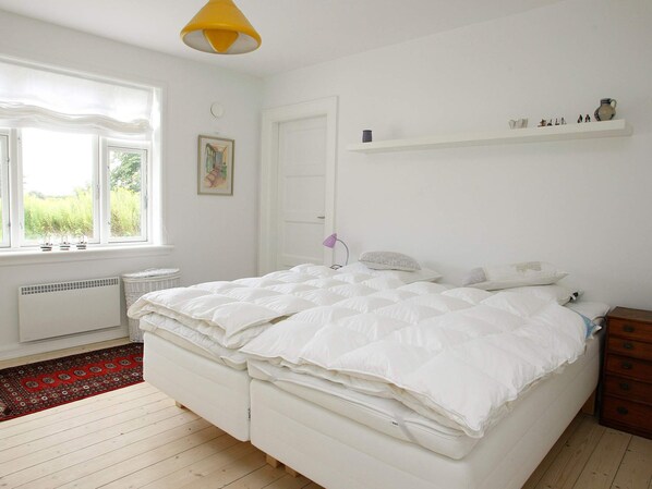 A bedroom with a bed, a window, a radiator, a shelf with decorative items, and a door.