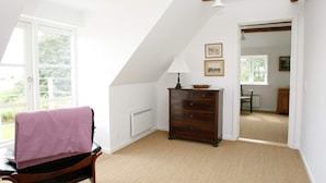 Una habitación con techo inclinado, una silla de madera con una toalla rosa, un aparador y una lámpara.