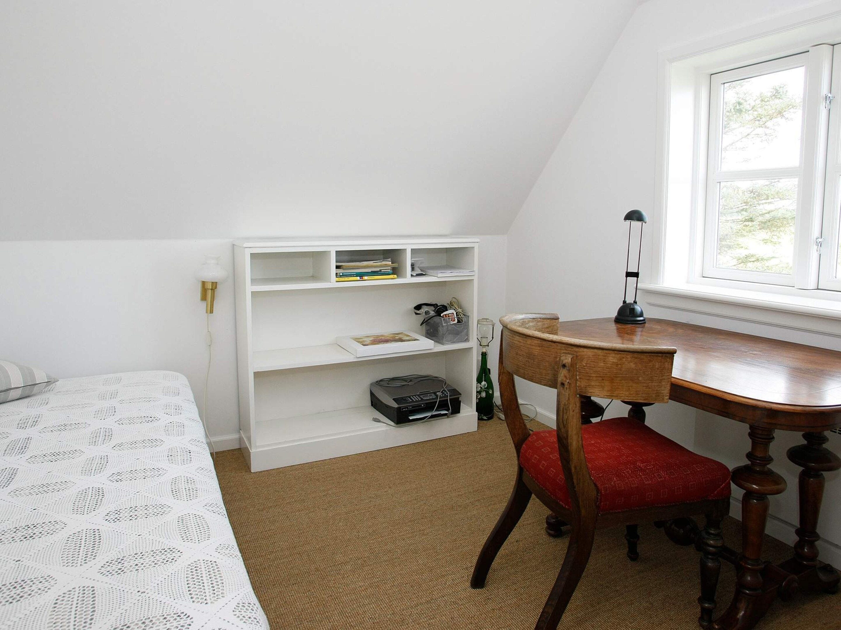 A room with a bed, a wooden table, a chair, a bookshelf, and a window.