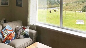 Quarto com uma grande janela que dá para um campo gramado com ovelhas. Há um sofá bege com almofadas coloridas e duas molduras com quadros na parede.
