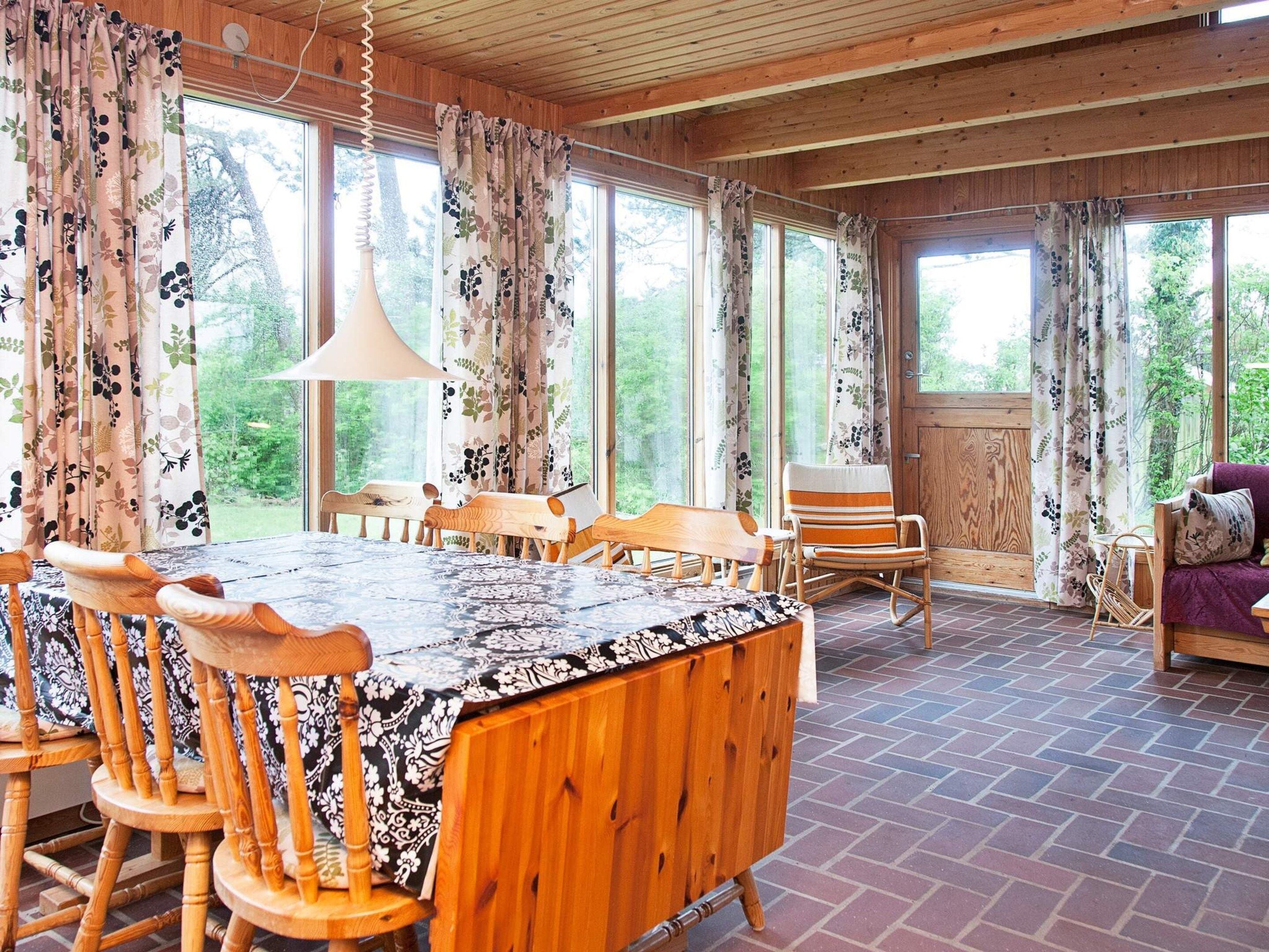A wooden dining area with a table and chairs, large windows with curtains, and a view of greenery outside.