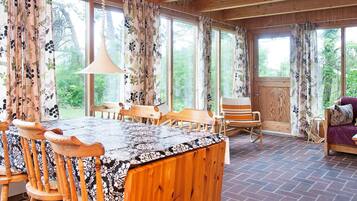 A wooden dining area with a table and chairs, large windows with curtains, and a view of greenery outside.