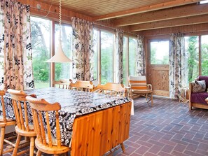 Un comedor de madera con mesa y sillas, ventanas amplias con cortinas, y vistas al exterior con vegetación.