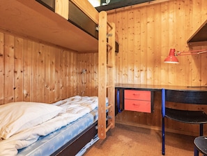 Quarto de madeira com uma cama de solteiro, uma escrivaninha com gavetas e uma luminária de mesa vermelha.