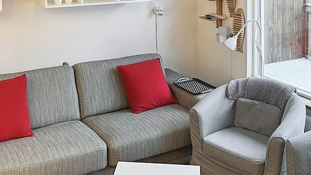 A living room with a grey sofa, red cushions, a patterned armchair, and a white coffee table. There are shelves with decorative items and a lamp on the wall. - Holiday Home in Lidköping (Lidköping)