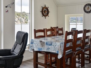 A dining area with a table covered in a patterned tablecloth, wooden chairs, a wall-mounted ship's wheel, and a clock. - Holiday Home in Urangsvåg (Bomlo)