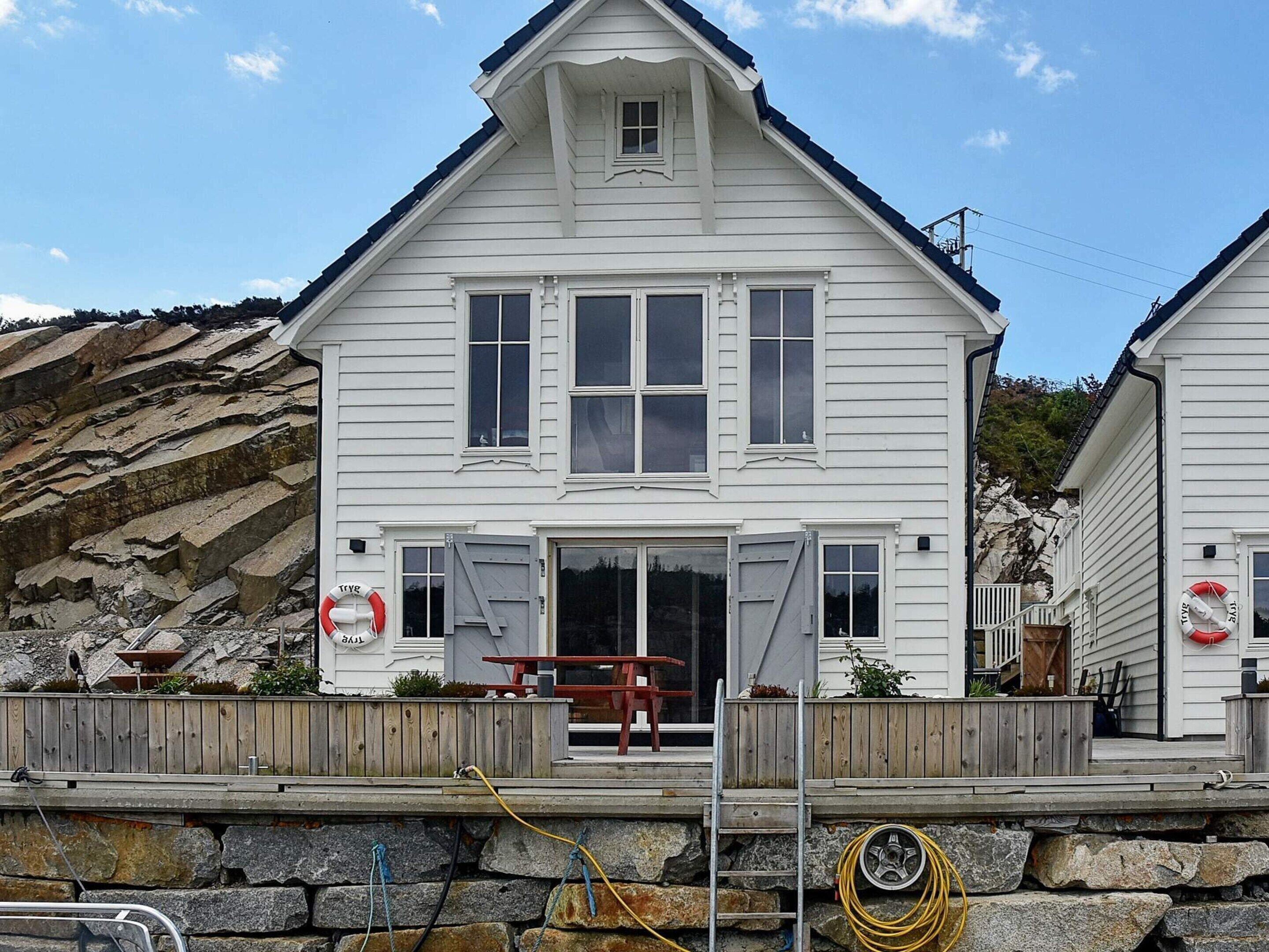 Een wit huis met twee verdiepingen, een voortuin, een houten terras en een stenen steunmuur.